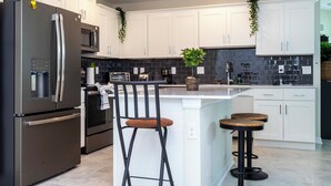 A modern kitchen with white cabinets, stainless steel appliances, a central island with stools, and decorative plants.