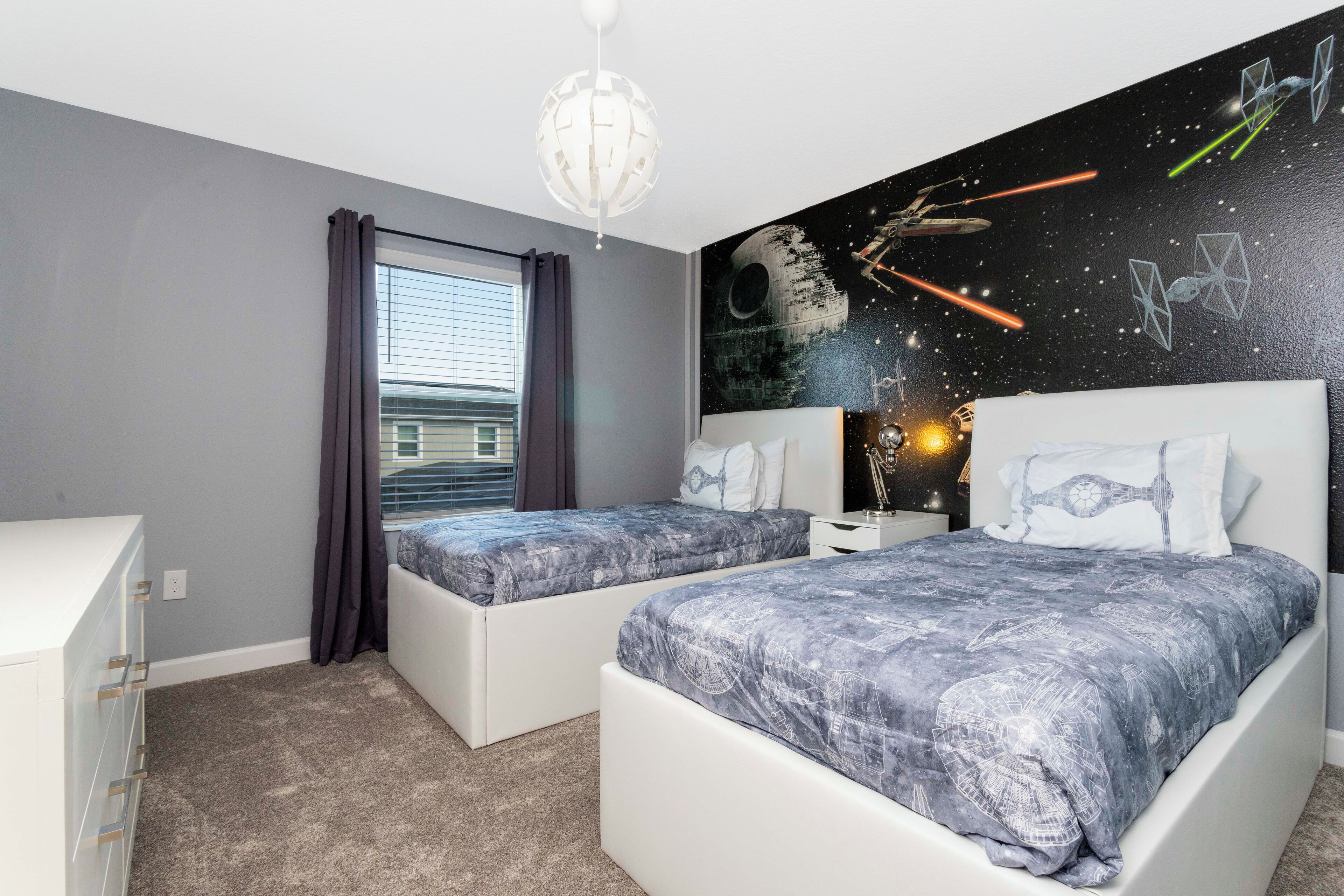 A bedroom with two beds, a dresser, a window with curtains, and a Star Wars-themed wall mural.