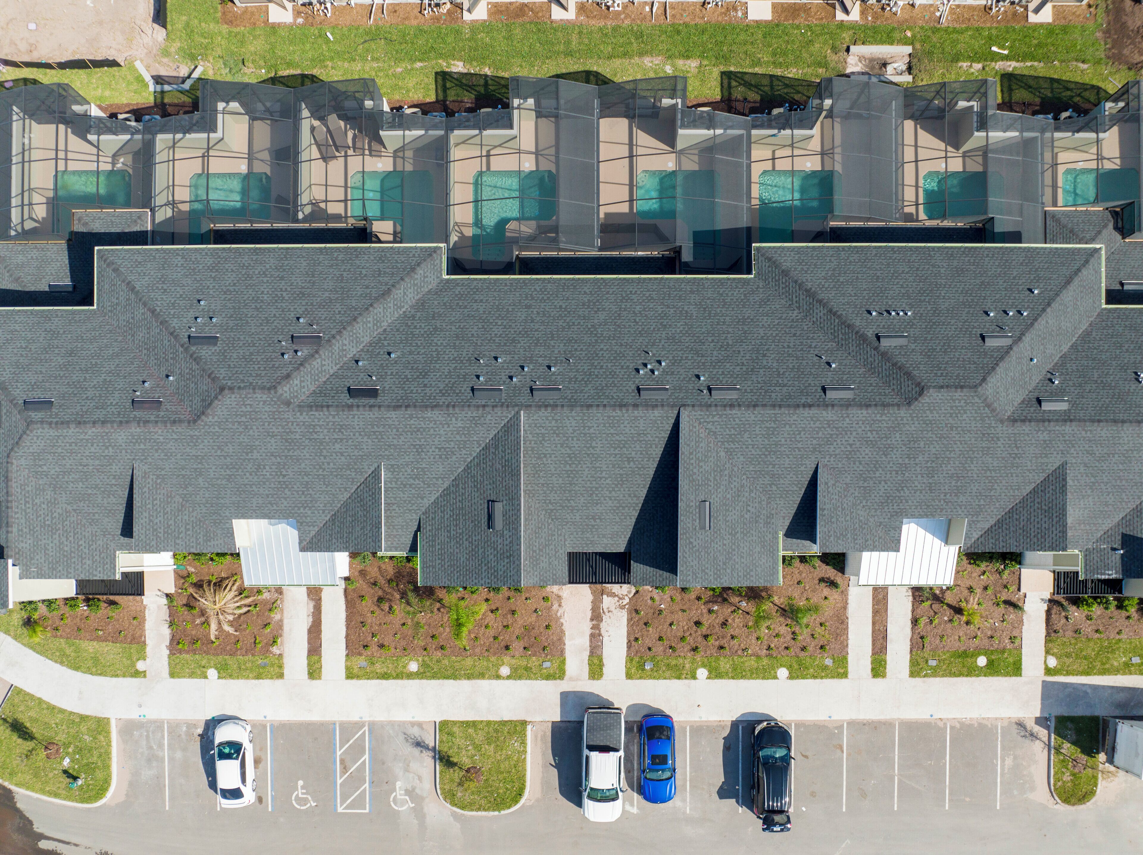 Une vue aérienne d’un parking où sont garées plusieurs voitures, avec une pelouse verdoyante et un bâtiment doté d’un toit au design géométrique original.
