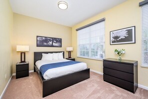 Un dormitorio con cama, mesitas de noche, cómoda, ventana con persianas y dos cuadros en la pared.