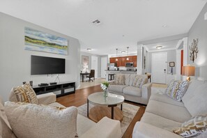 A modern living room with a flat-screen TV, comfortable seating, a coffee table, and a kitchen in the background. - Epic Universe NEW Beautiful Lakeview Condo Near the Parks - 1026 (Orlando)