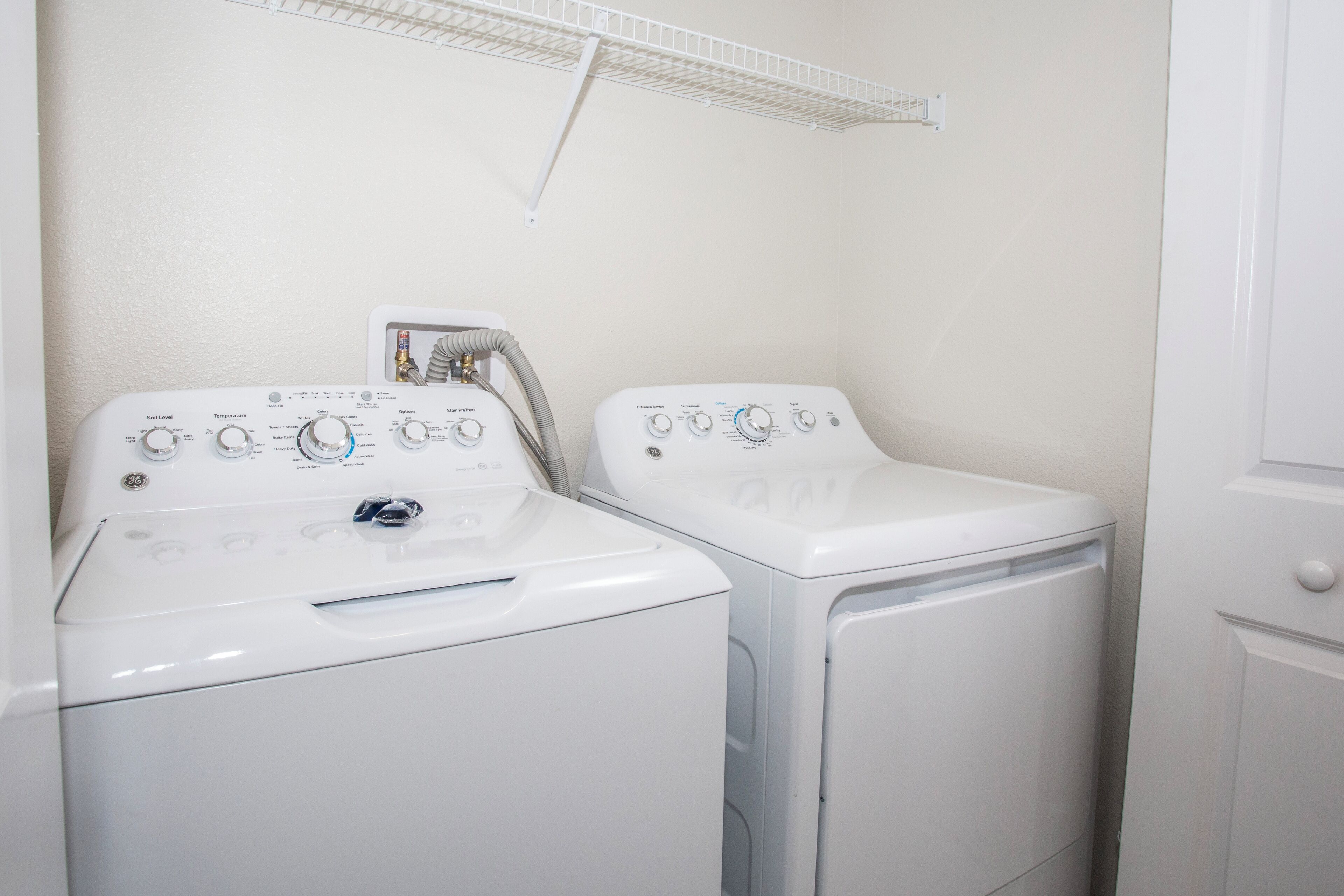 Two front-loading washing machines in a laundry room with a white wall and a metal shelf above.