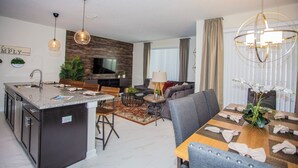 A modern kitchen with a central island, dining area with a table set for four, and a living room with a sofa and a TV. - Four Bedroom Storey Lake Resort Close to Disney w Pool 3063 (Kissimmee)