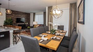 A modern dining area with a wooden table set for six, surrounded by upholstered chairs. A large chandelier hangs above the table, and a television is mounted on the wall to the left. - Four Bedroom Storey Lake Resort Close to Disney w Pool 3063 (Kissimmee)