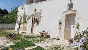 Un edificio blanco con una escalera de piedra que lleva a una puerta, un pequeño jardín con una mesa y sillas, y una planta en maceta.