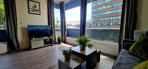 A modern living room with a large flat-screen TV, a sofa, a coffee table, and a balcony with potted plants. - Inviting 1-bed Apartment in Hemel Hempstead (Hemel Hempstead)