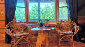 Een gezellig chaletinterieur met twee rieten stoelen rond een houten tafel, een raam met gordijnen en uitzicht op groen buiten.