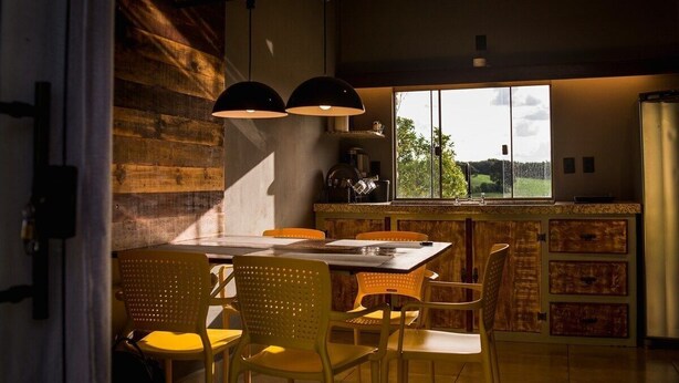 A dining area with a wooden table and chairs, a window with a view of greenery, and pendant lights. - Farmless Home (Tiradentes)