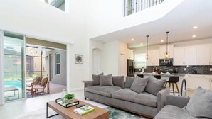 A modern living room with a grey sofa, a coffee table, and a view of the kitchen and outdoor area. - Gorgeous 5Bd Single Family w Pool Storey Lake 2767o (Kissimmee)