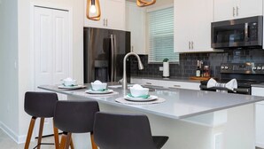 A modern kitchen with white cabinets, a stainless steel refrigerator, a black microwave, and a central island with bar stools. - Gorgeous 5Bd Single Family w Pool Storey Lake 2767o (Kissimmee)