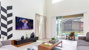 A modern living room with a large TV, a sofa, a coffee table, and a view of a pool through large windows. - Gorgeous 5Bd Single Family w Pool Storey Lake 2767o (Kissimmee)
