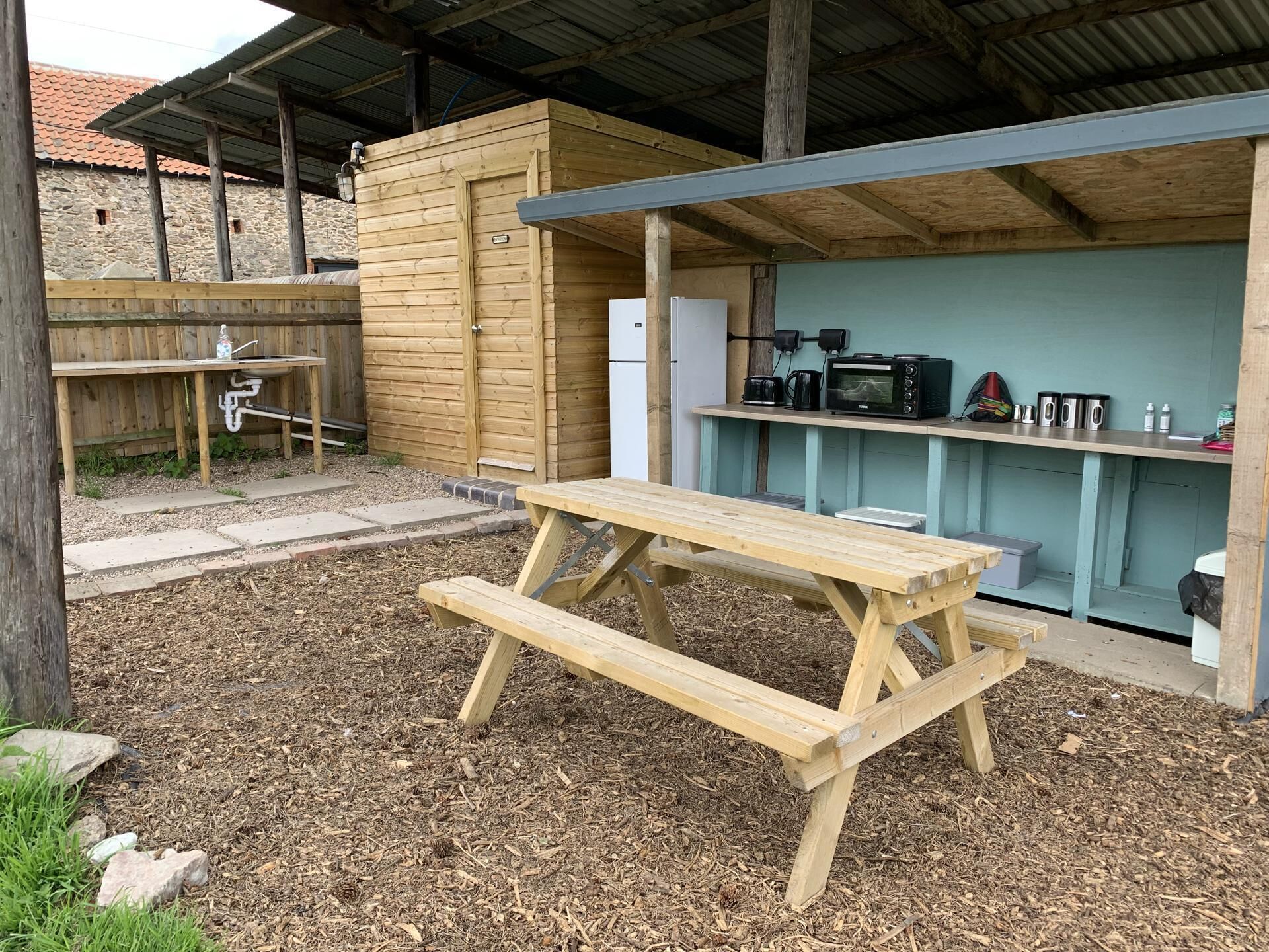 Una mesa de picnic de madera en un espacio exterior cubierto, con un muro de piedra y techo de metal.