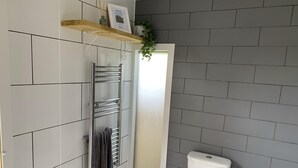 A modern bathroom with grey tiled walls, a white toilet, a towel rack, and a shelf with a picture and a plant. - Double Bell Tent Farm Stay (Loughborough)