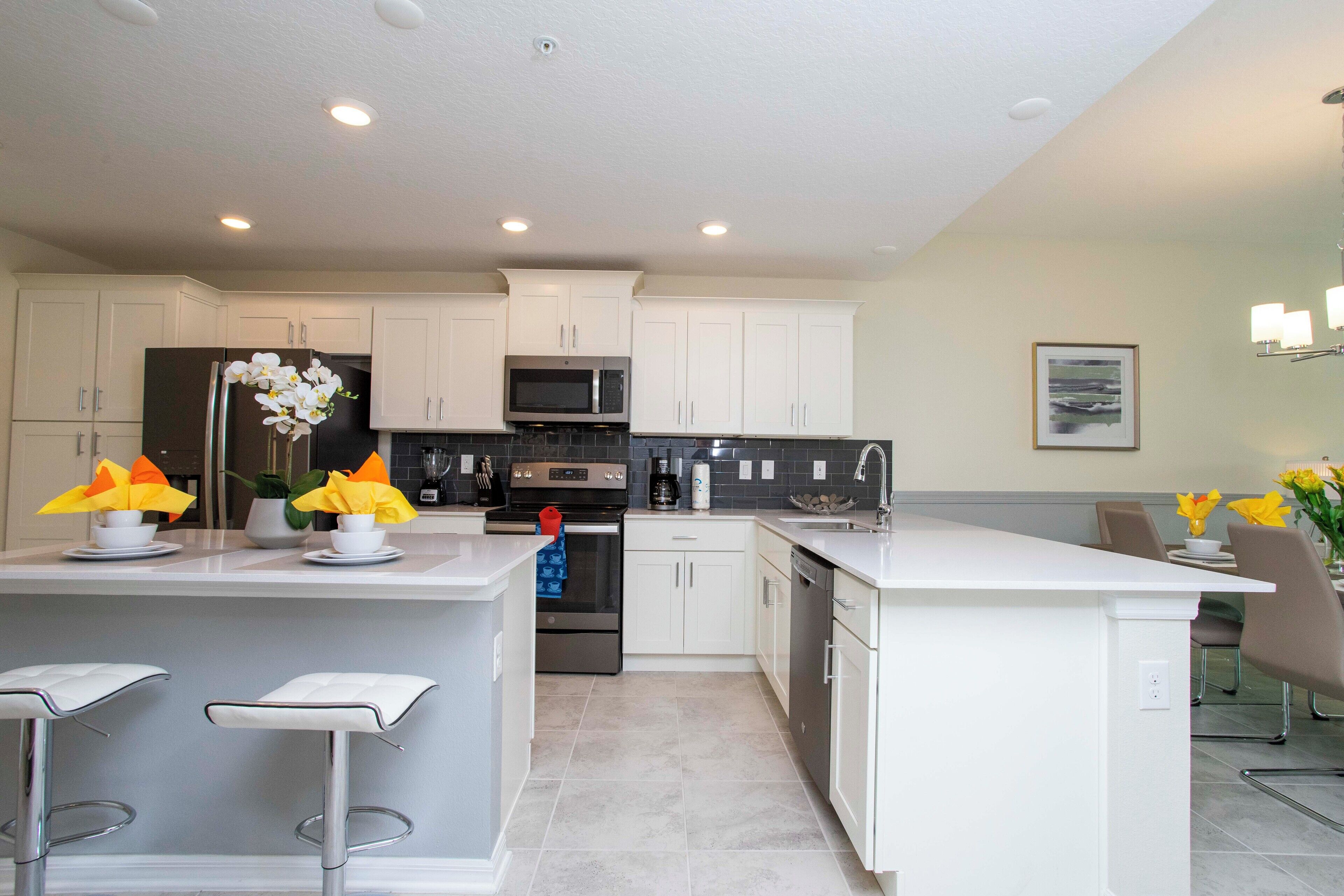 A modern kitchen with white cabinets, a central island with bar stools, stainless steel appliances, and decorative flowers.