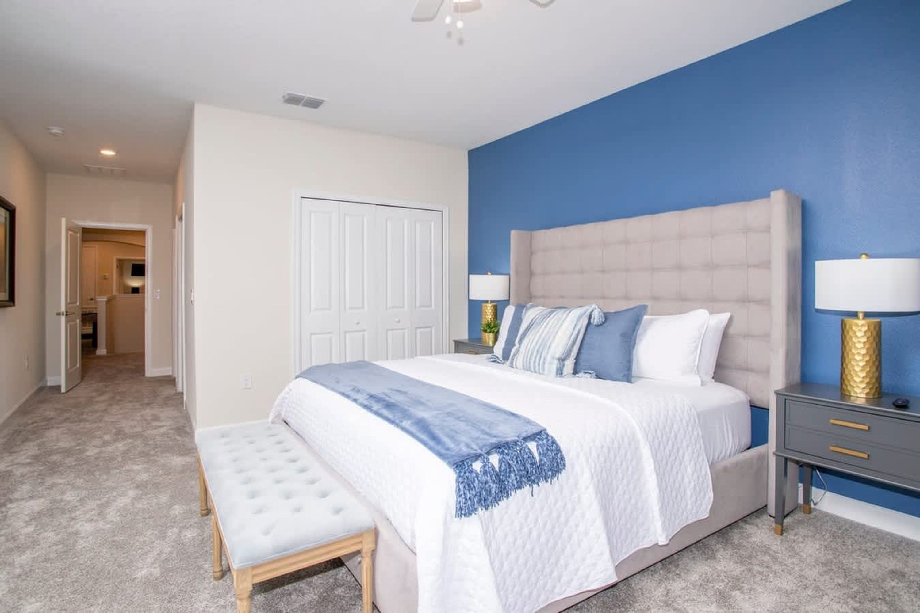 A bedroom with a large bed, bedside tables, a bench, a door, and a hallway.