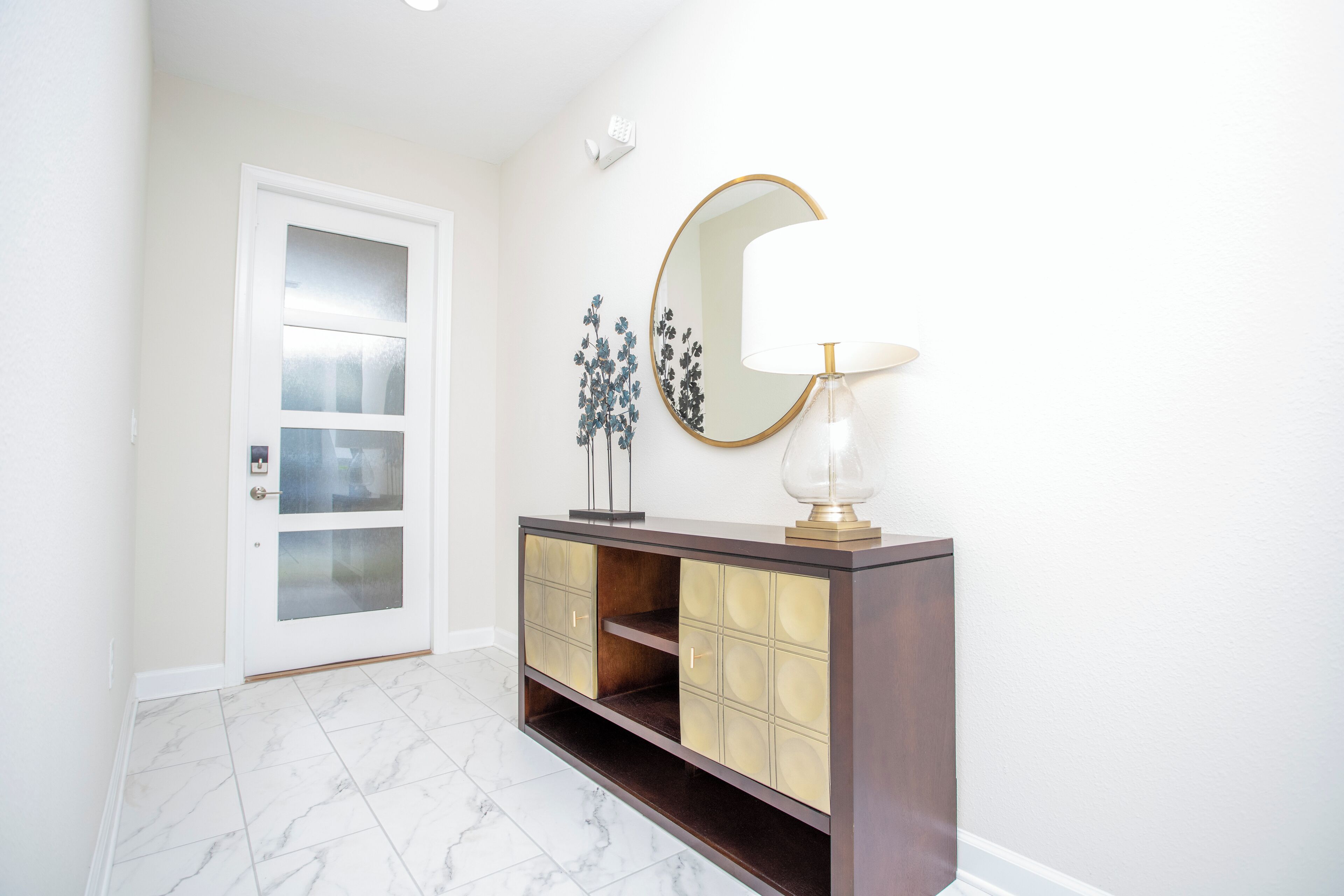 A hallway with a white door, a dark wooden console with glass doors, a round mirror, and a table lamp.