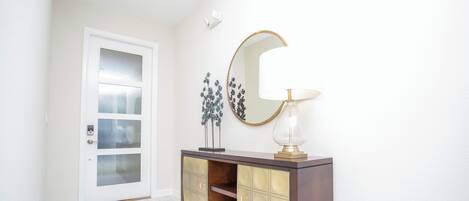 A hallway with a white door, a dark wooden console with glass doors, a round mirror, and a table lamp.