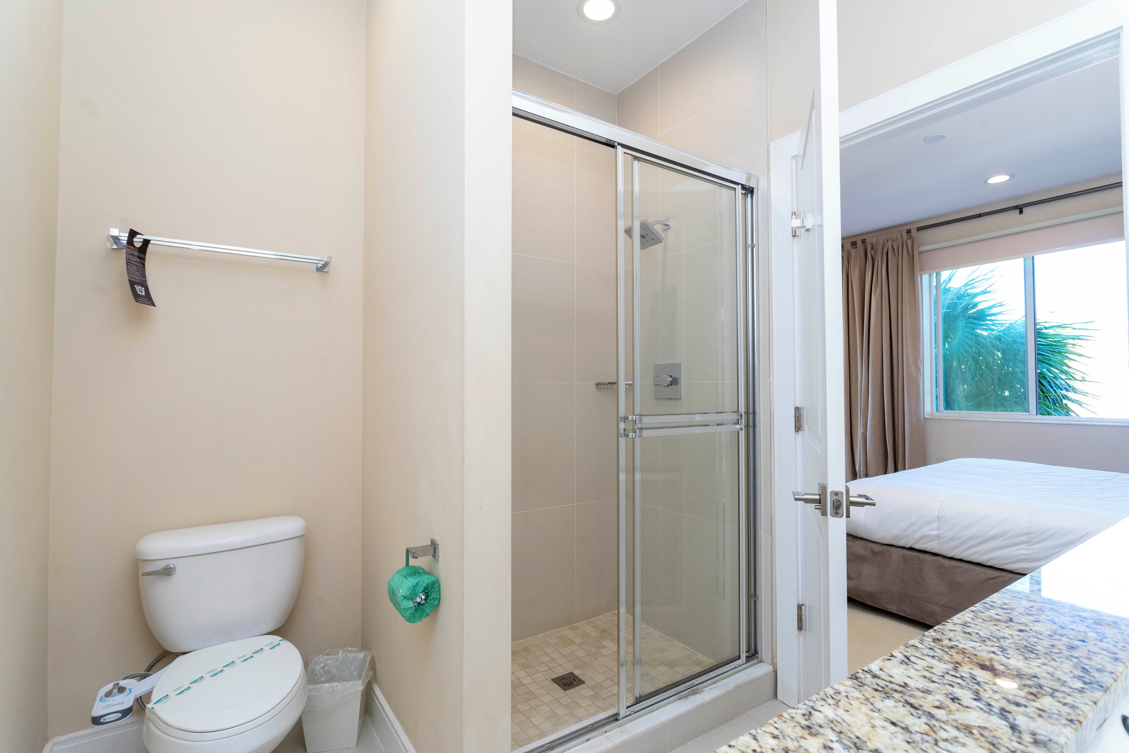 A bathroom with a toilet, a glass shower enclosure, and a bed in the adjacent room.
