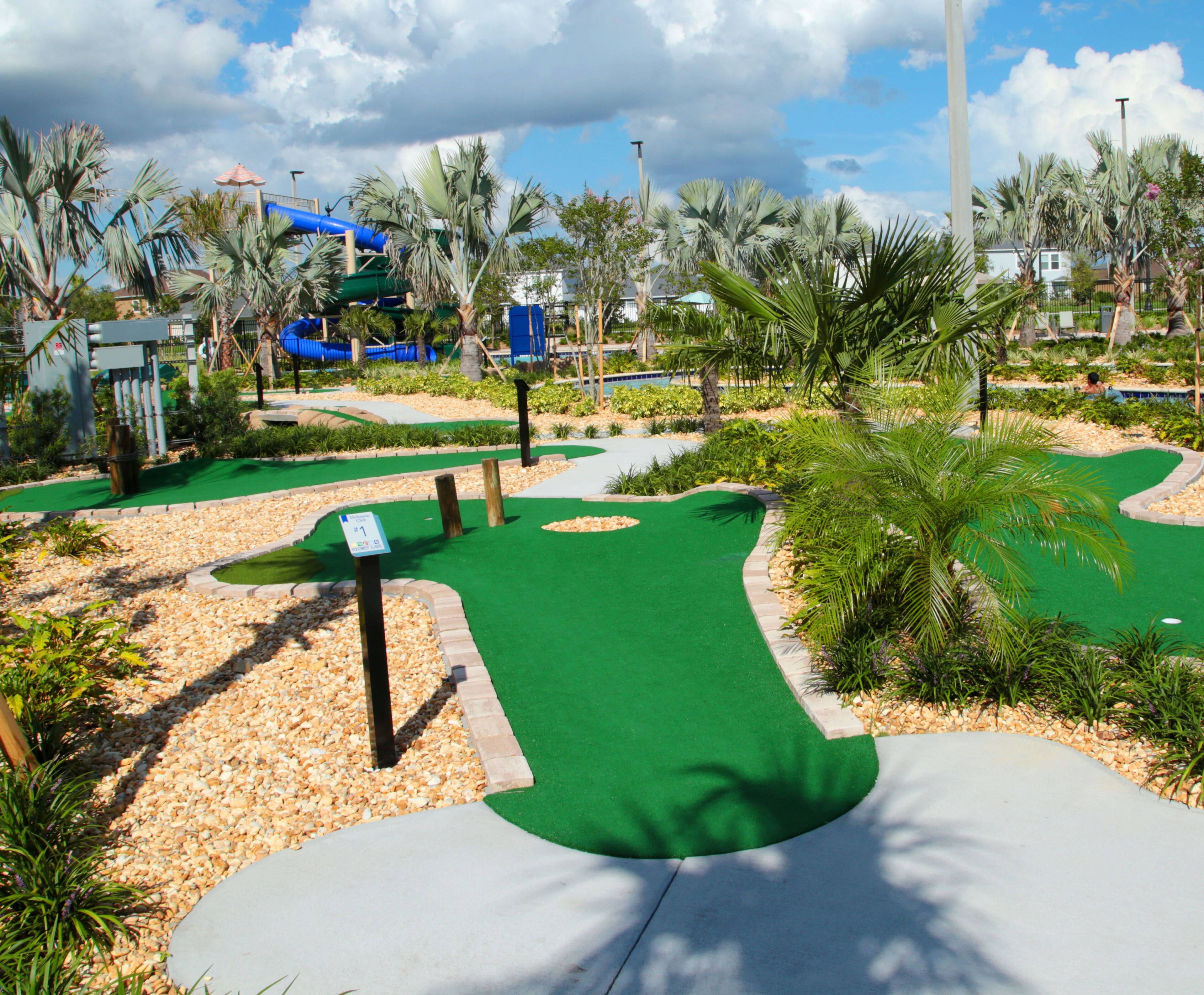Un campo de mini golf con superficie de césped verde, palmeras y un tobogán de agua azul al fondo.