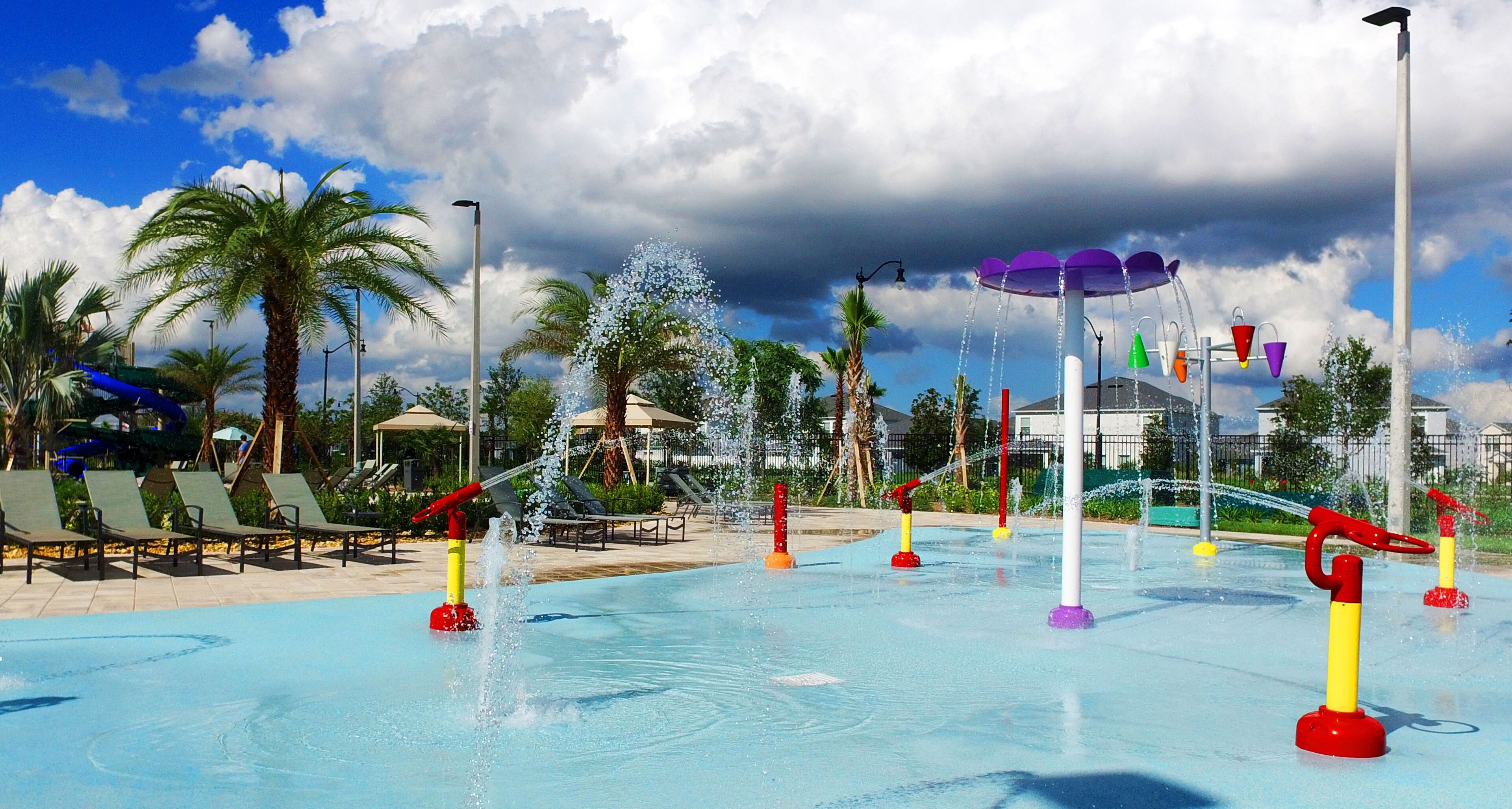 A water park with multiple water features, including fountains and slides, surrounded by palm trees and lounge chairs.