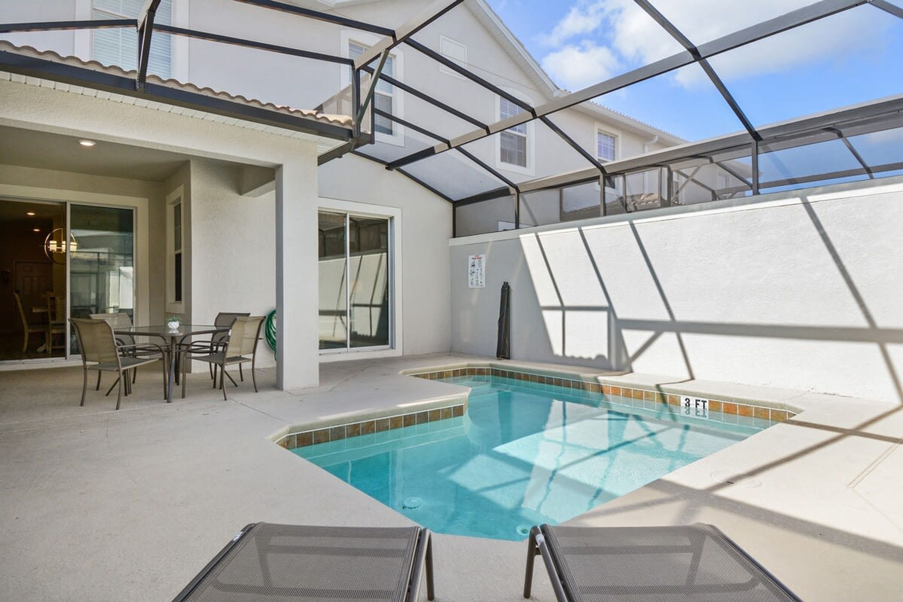 A covered outdoor swimming pool with a glass enclosure, a patio table, and chairs.