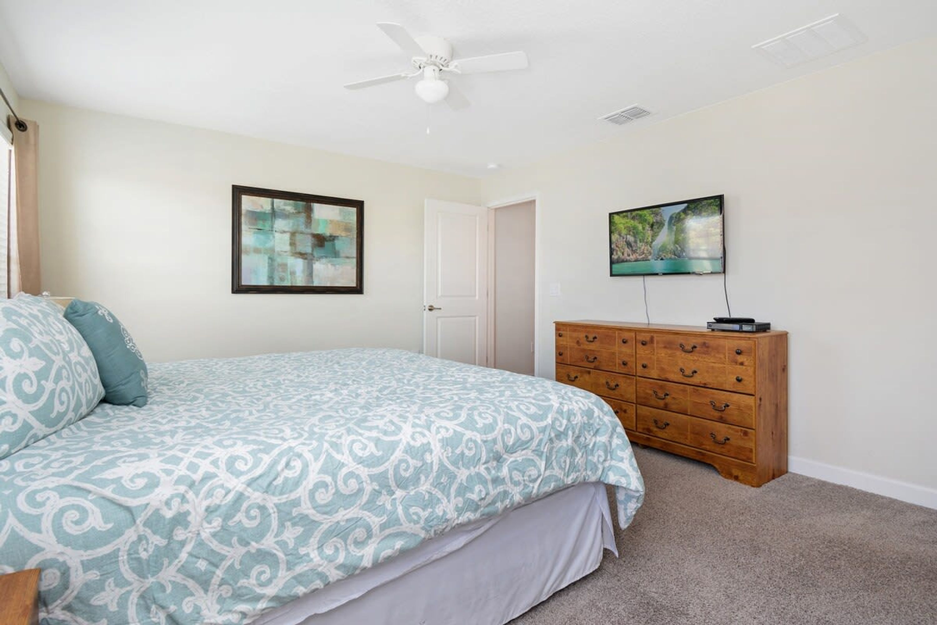 Une chambre à coucher avec un lit, une commode en bois, un ventilateur de plafond et deux tableaux encadrés accrochés au mur.