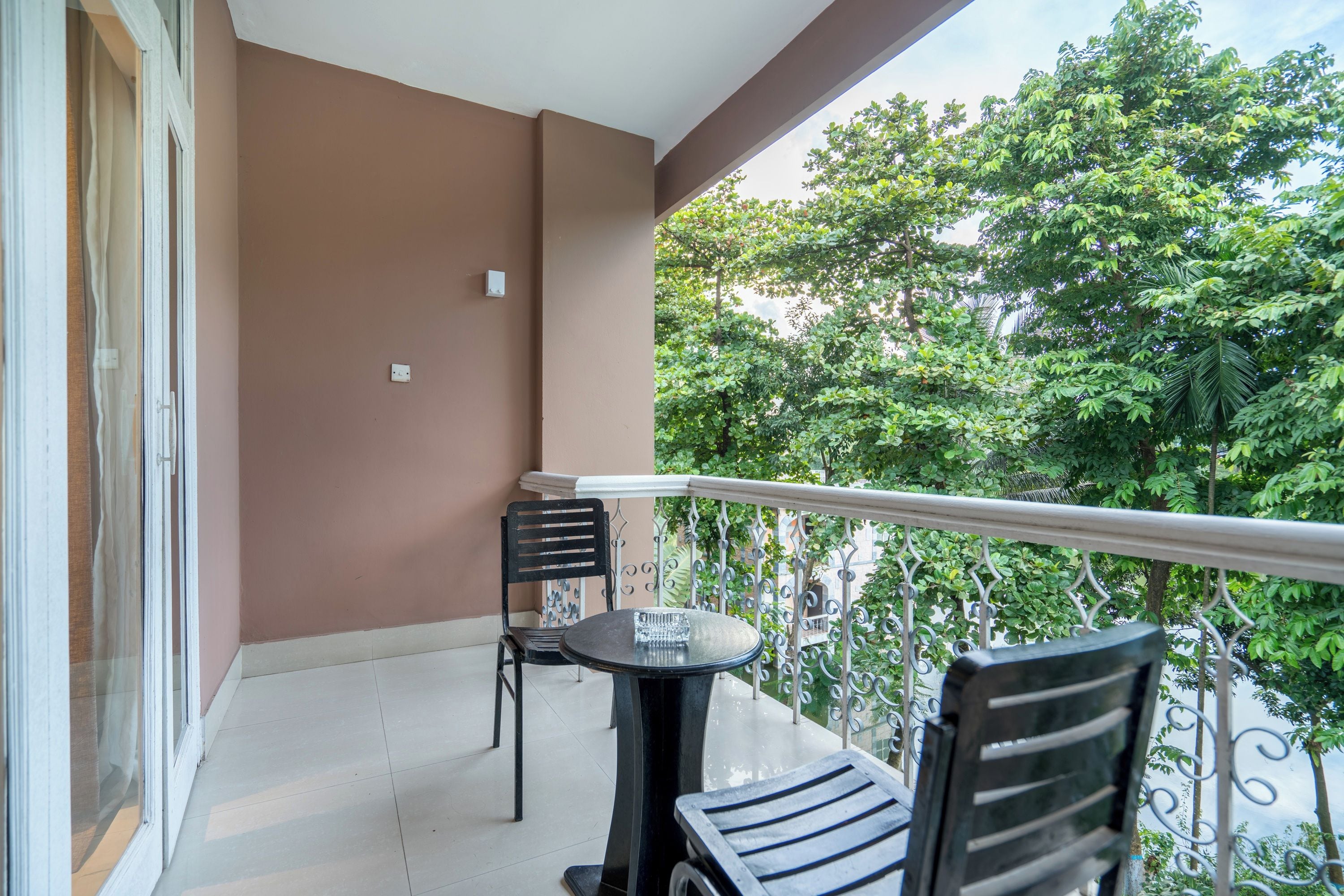 A balcony with a table and two chairs, a view of trees, and a brown wall.