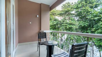 A balcony with a table and two chairs, a view of trees, and a brown wall.