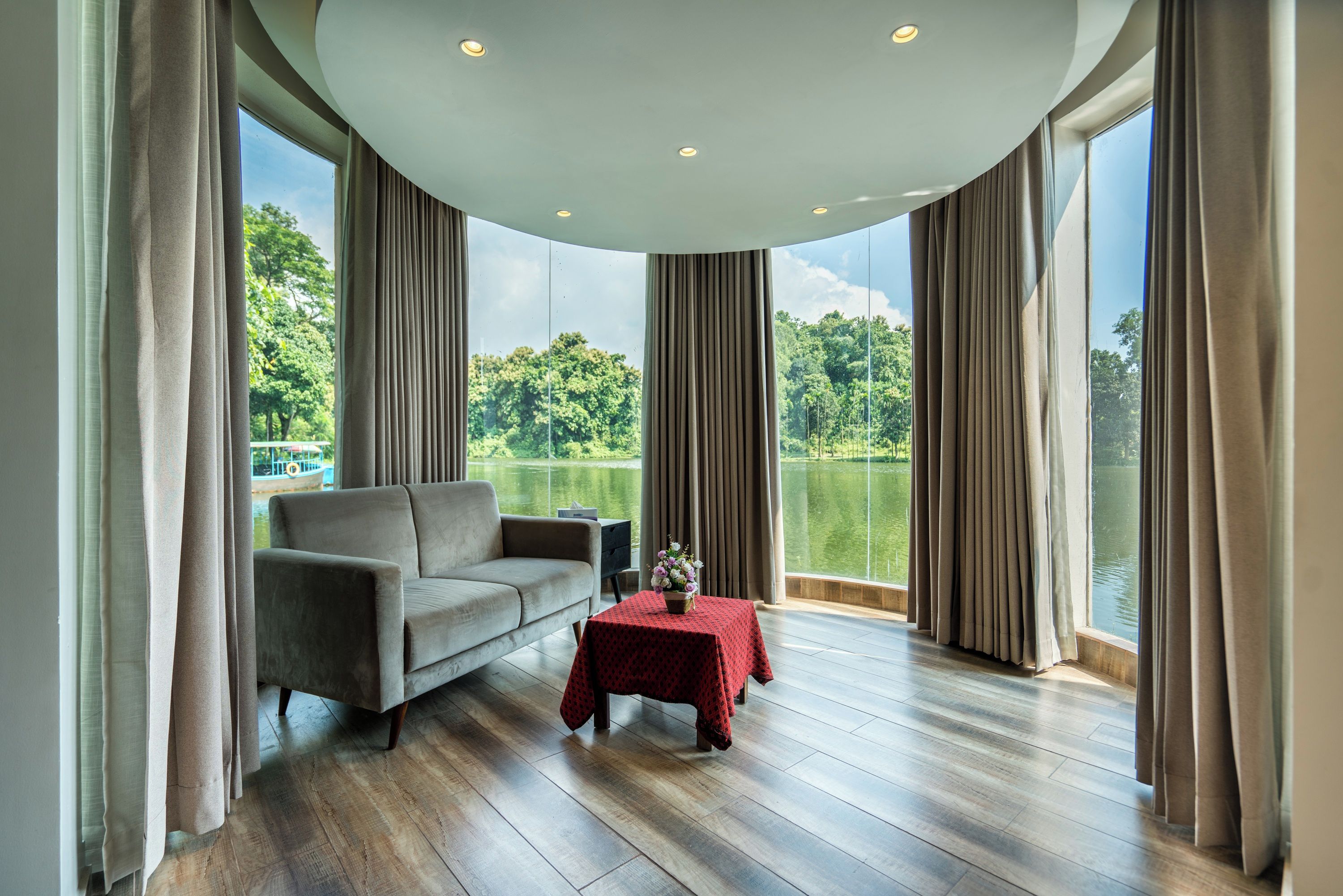 A room with a sofa, a small table with a red tablecloth, and a view of a lake through large windows.