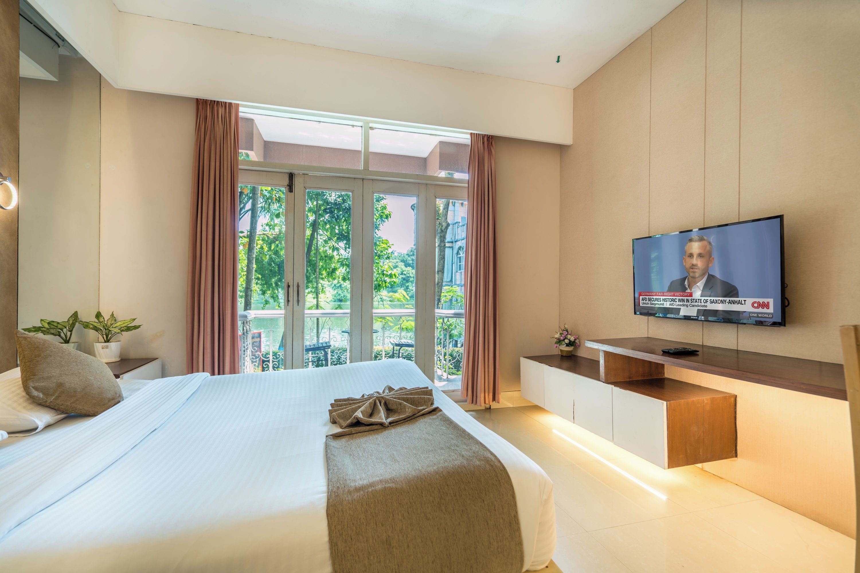 Habitación de hotel con cama, televisión en la pared, balcón con cortinas y una pequeña mesa con una planta.