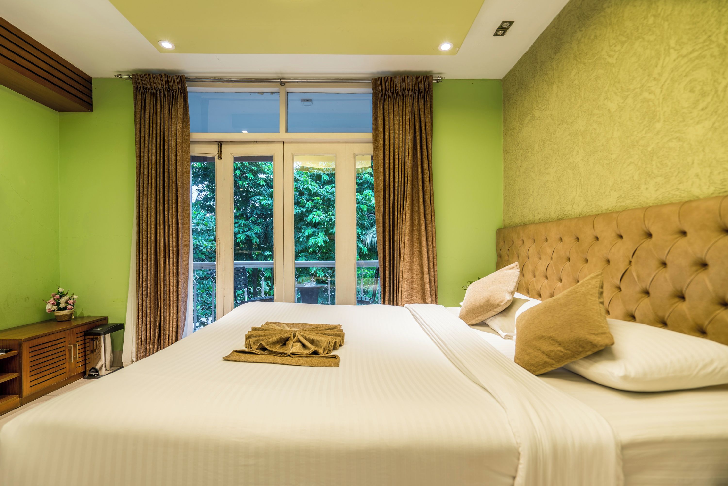 A hotel room with a large bed, a view of greenery through the window, and a wooden nightstand.