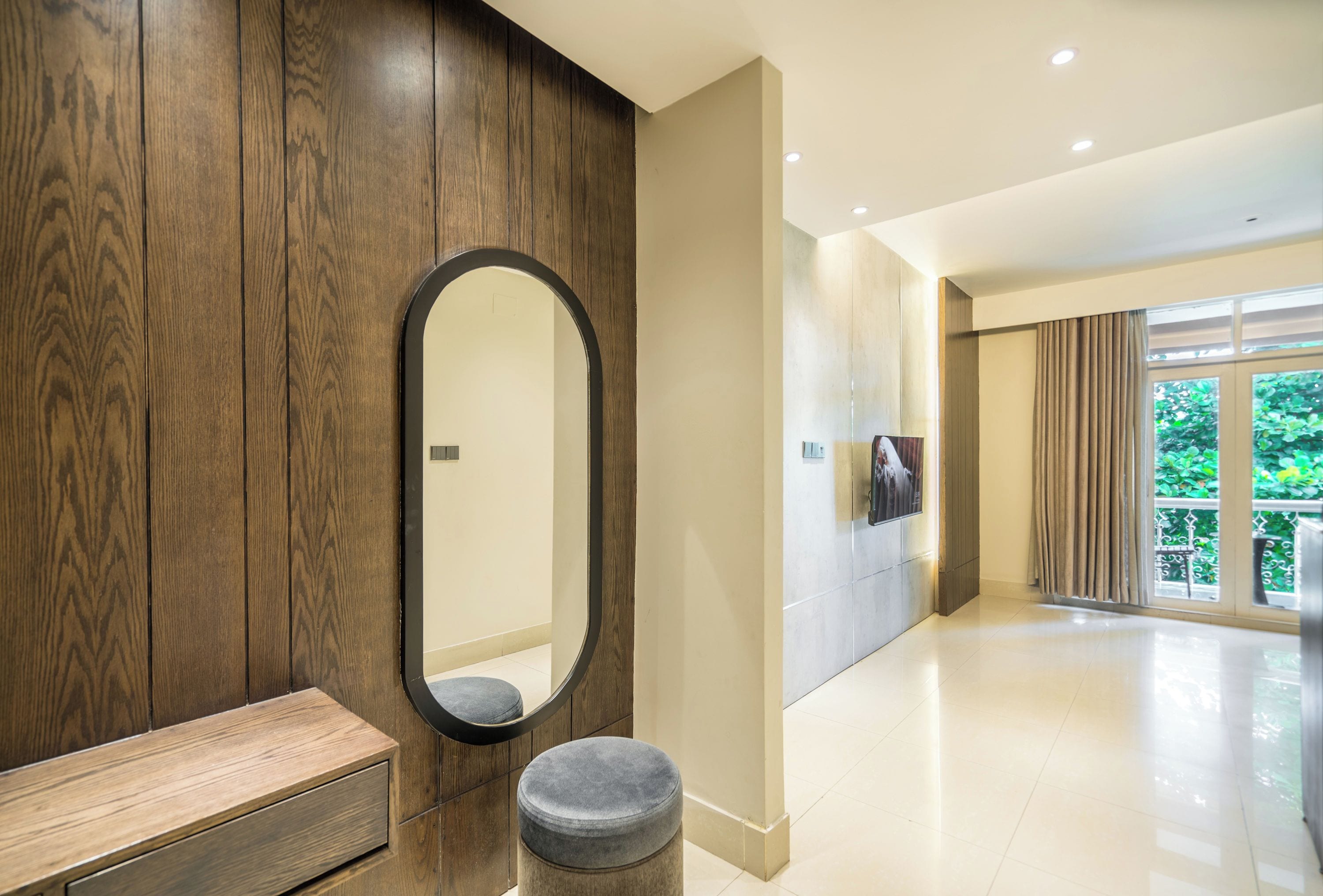 A modern bathroom with a wooden vanity, a round mirror, and a grey ottoman.