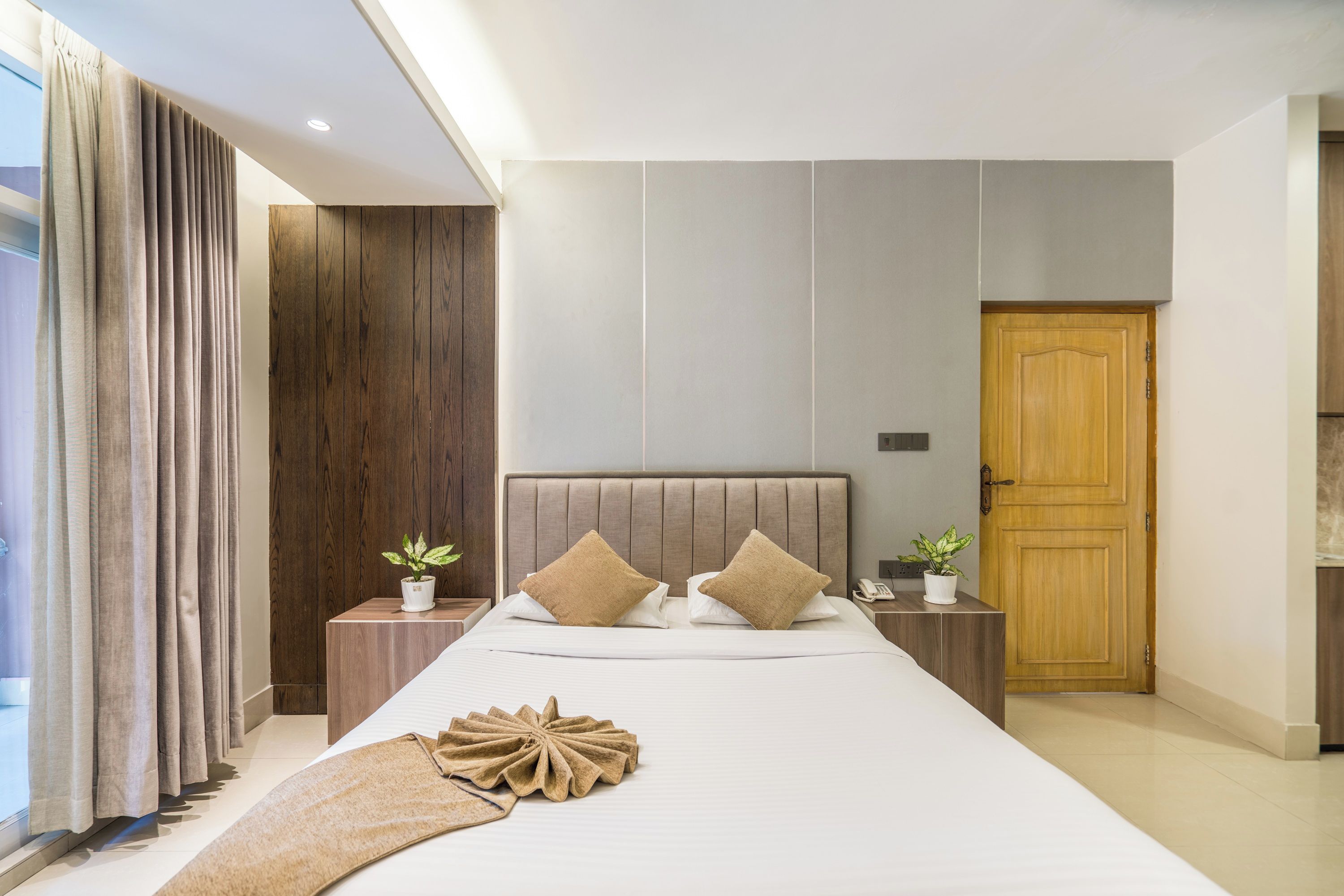 Een moderne hotelkamer met een groot bed, een houten hoofdeinde, twee nachtkastjes met planten, een houten deur en een groot raam met gordijnen.