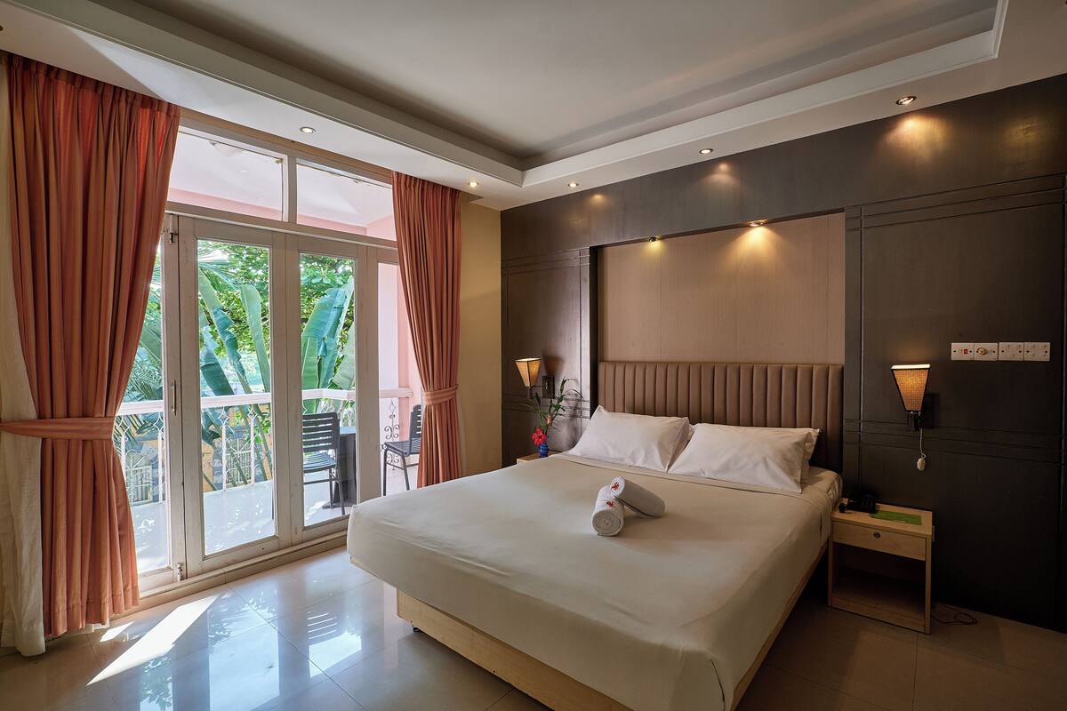 A hotel room with a large bed, a nightstand, a wall-mounted lamp, and a sliding glass door leading to an outdoor area.
