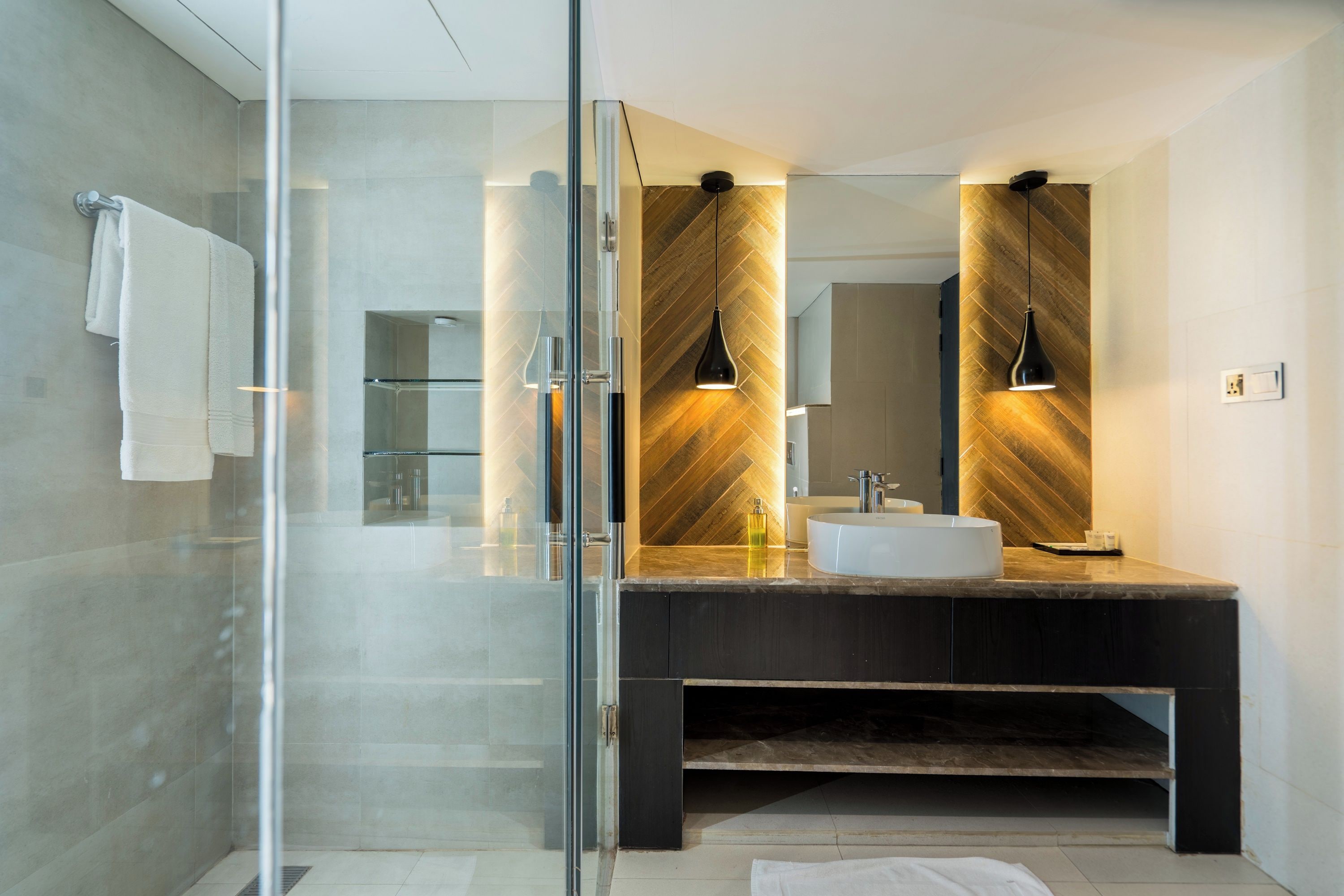Une salle de bain moderne avec une douche à l’italienne en verre, un grand miroir et un meuble-lavabo foncé avec un vasque blanche.