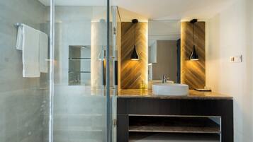 Une salle de bain moderne avec une douche à l’italienne en verre, un grand miroir et un meuble-lavabo foncé avec un vasque blanche.