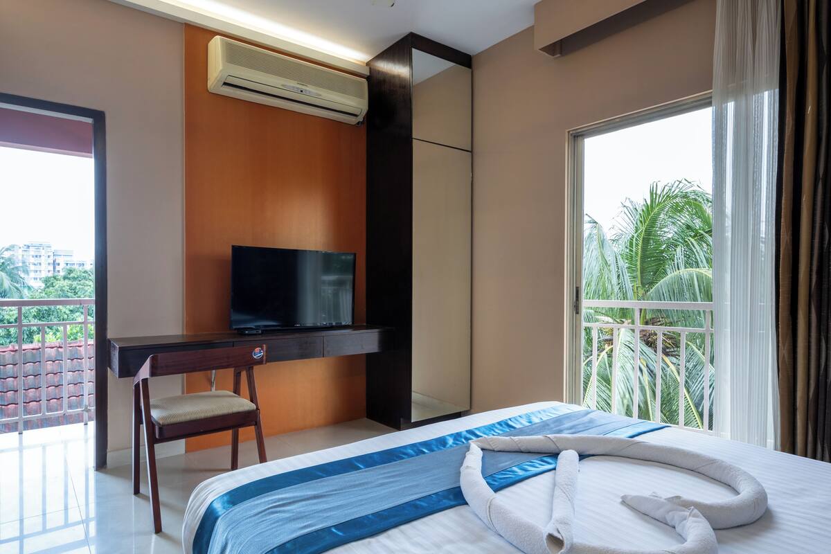 A hotel room with a bed, a desk, a television, and a balcony with a view of palm trees.