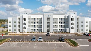 Un hotel de varios pisos con un diseño moderno, que cuenta con un patio central con una fuente, espacios para estacionamiento y un cielo azul despejado.