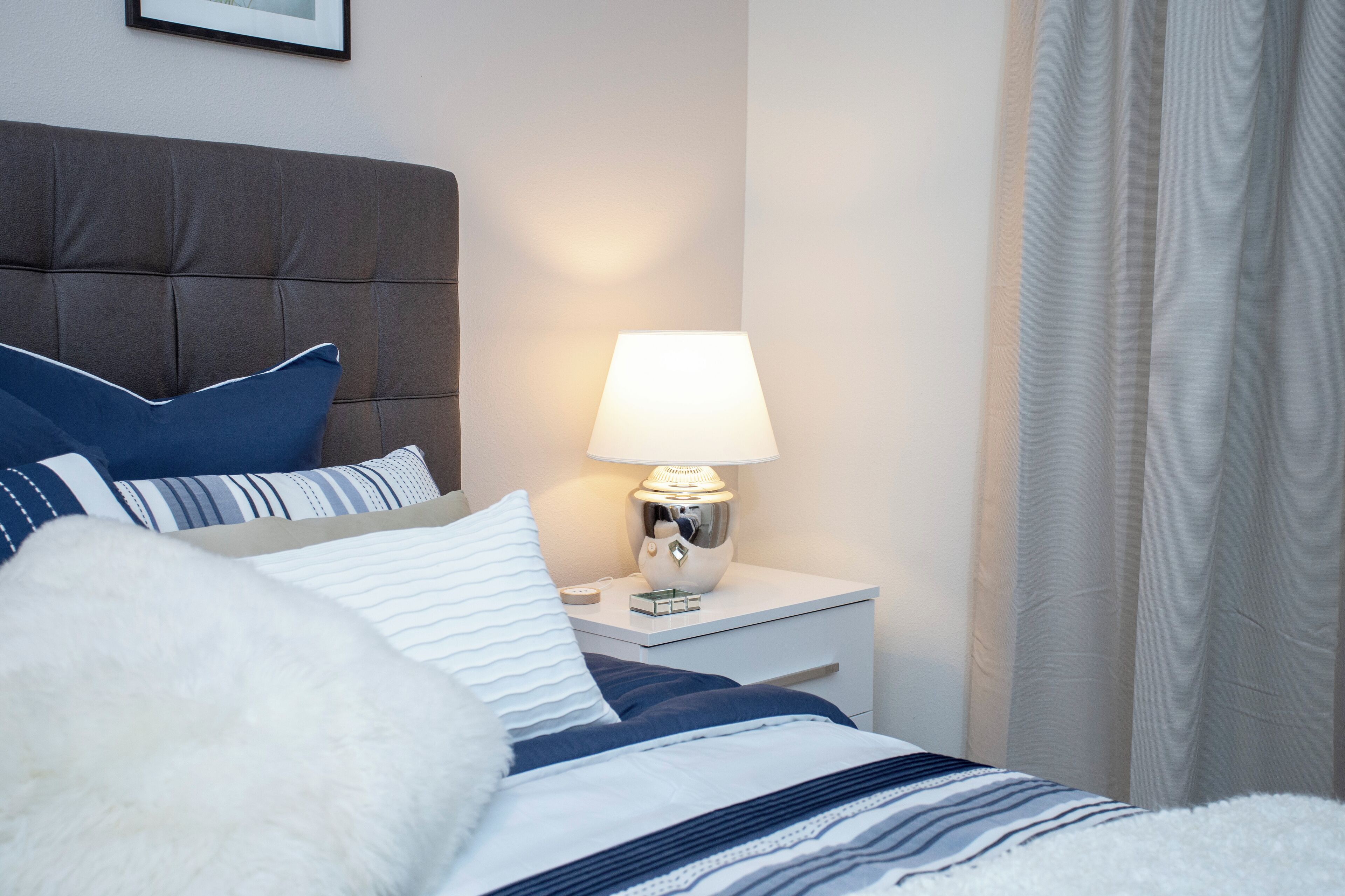 A neatly made bed with a striped duvet, a white fluffy pillow, and a blue pillow. There is a white bedside table with a lamp and a small clock. The room has a neutral color scheme with a grey headboard and white walls.