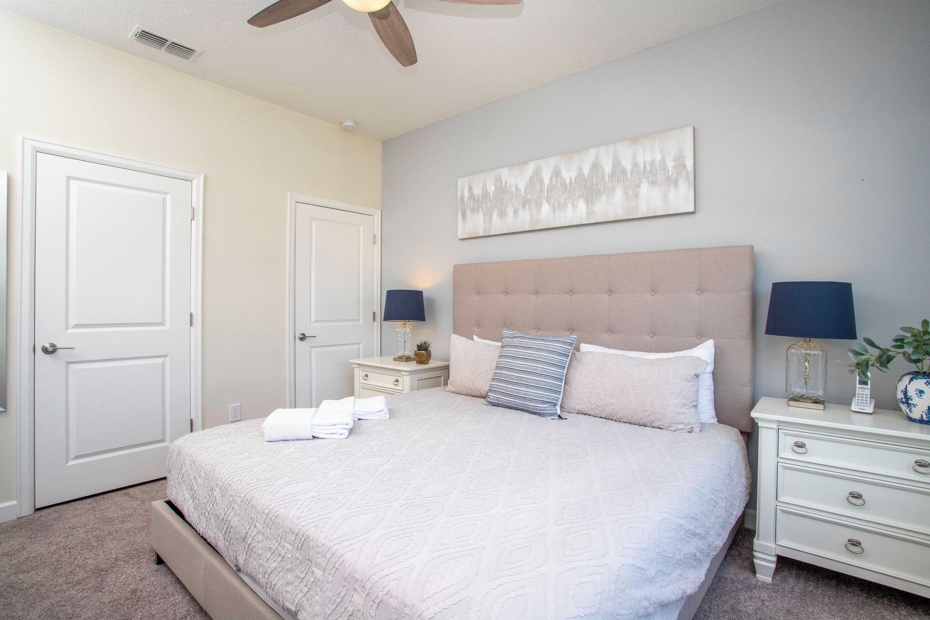 A bedroom with a large bed, two bedside tables, a ceiling fan, and a wall-mounted artwork.