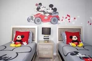 Ein Schlafzimmer mit zwei Betten, beide mit Bettdecken im Mickey-Maus-Design, einem Nachttisch mit Lampe und einer Wandsticker-Motiv von Mickey und Minnie Maus in einem Auto.