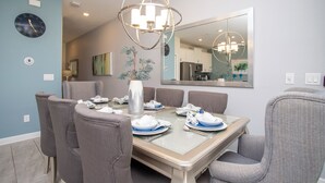 Uma sala de jantar com uma mesa posta para seis pessoas, rodeada por cadeiras estofadas cinzentas. Um lustre moderno pendura-se acima da mesa, e um espelho grande reflete o espaço.