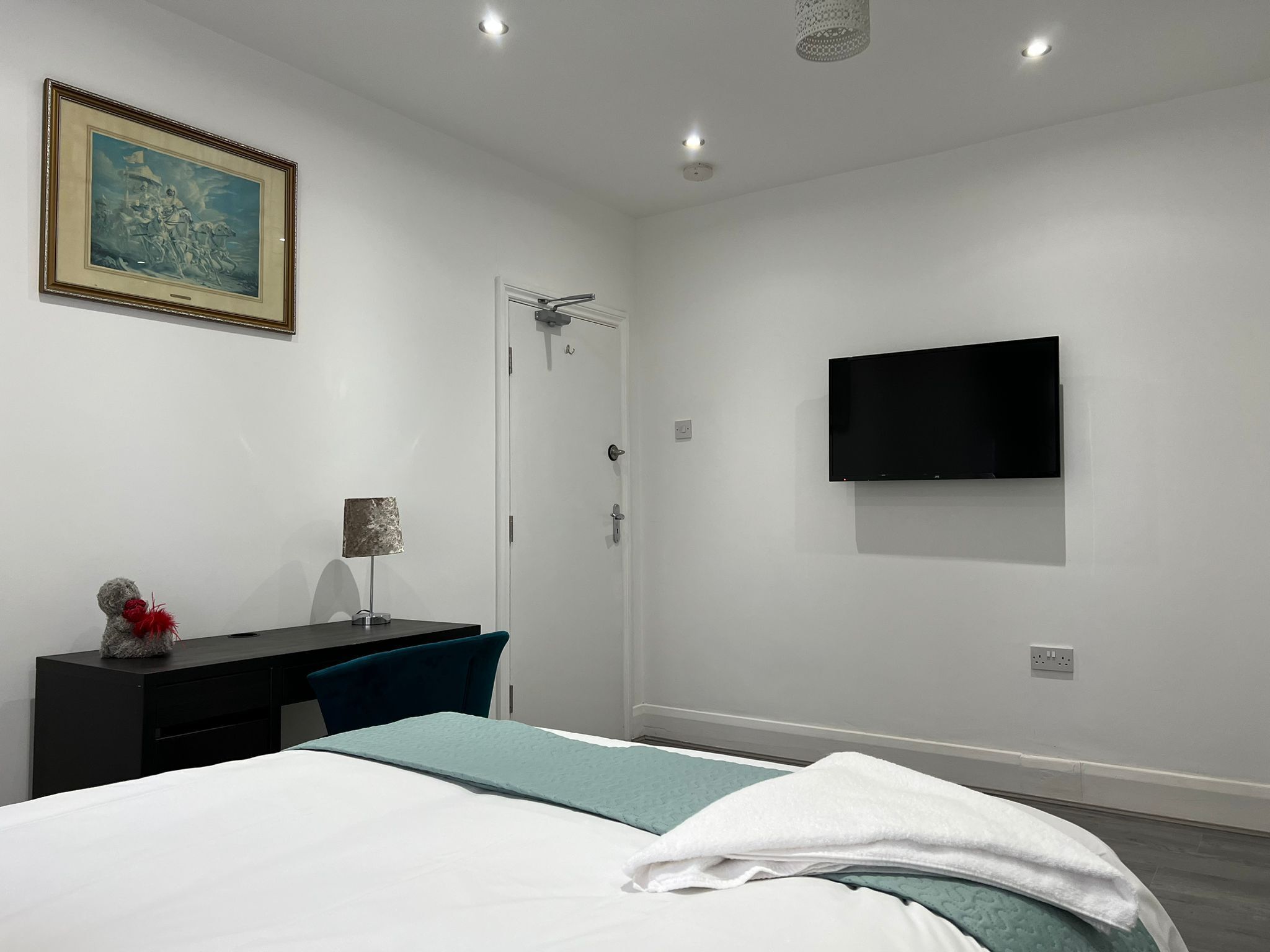 Ein modernes Hotelzimmer mit einem Bett, einem Schreibtisch mit Stuhl, einem an der Wand befestigten Fernseher und einer Tür.
