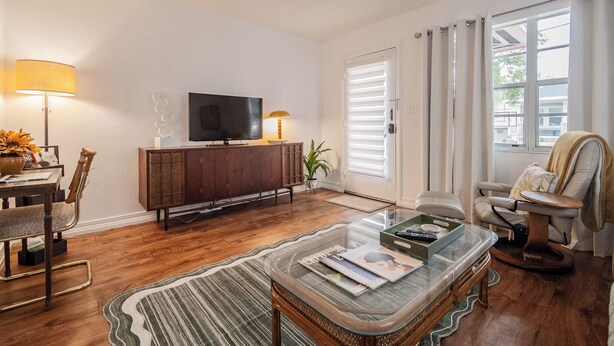 A living room with a wooden floor, a glass coffee table, a TV on a wooden cabinet, a dining area with a table and chairs, and a large window with curtains.