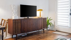 A modern living room with a flat-screen TV on a wooden cabinet, a yellow lamp, a potted plant, and a sliding glass door with blinds.