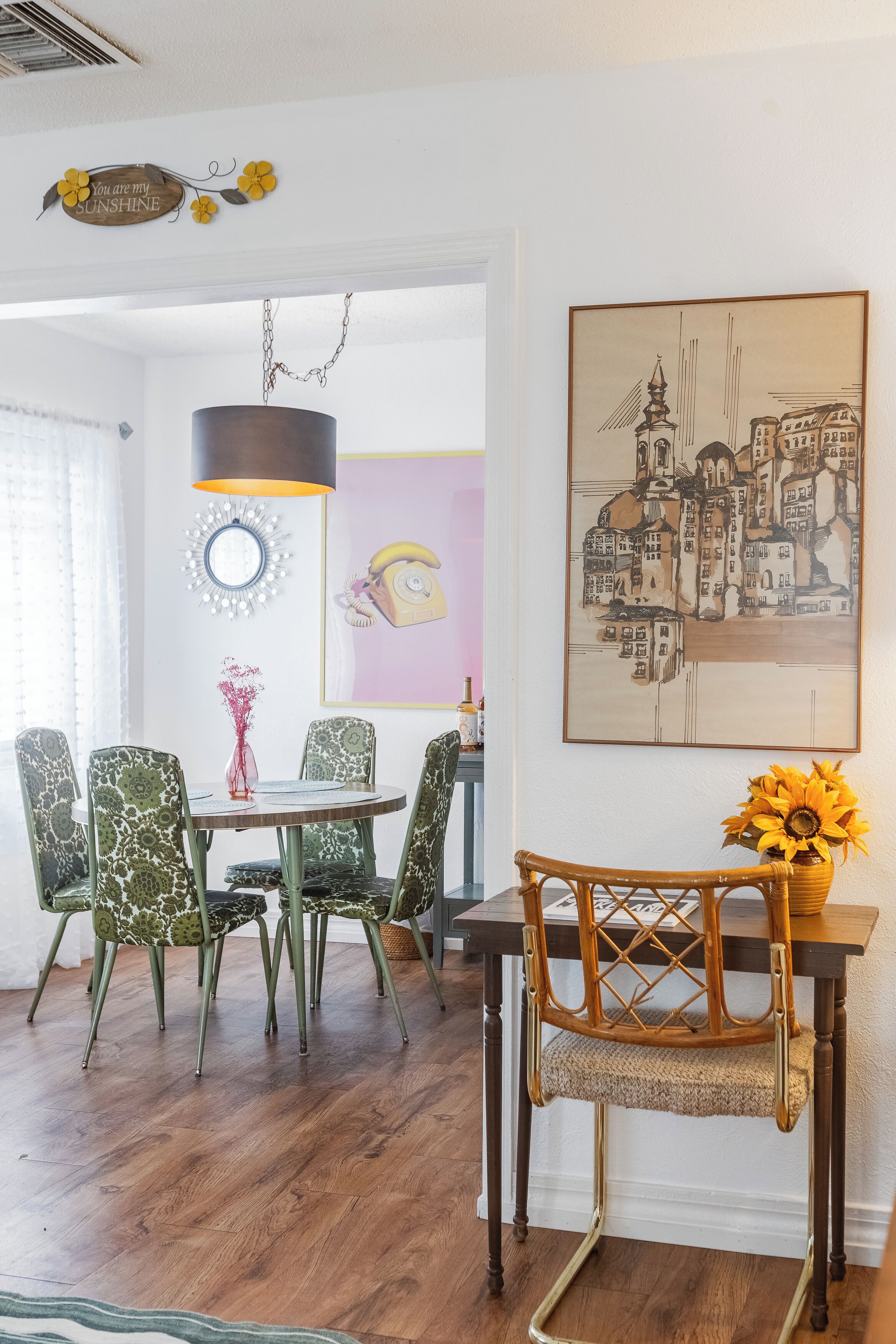 A dining area with a round table, patterned chairs, a wooden chair with a woven seat, a hanging lamp, a mirror, and a cityscape painting.