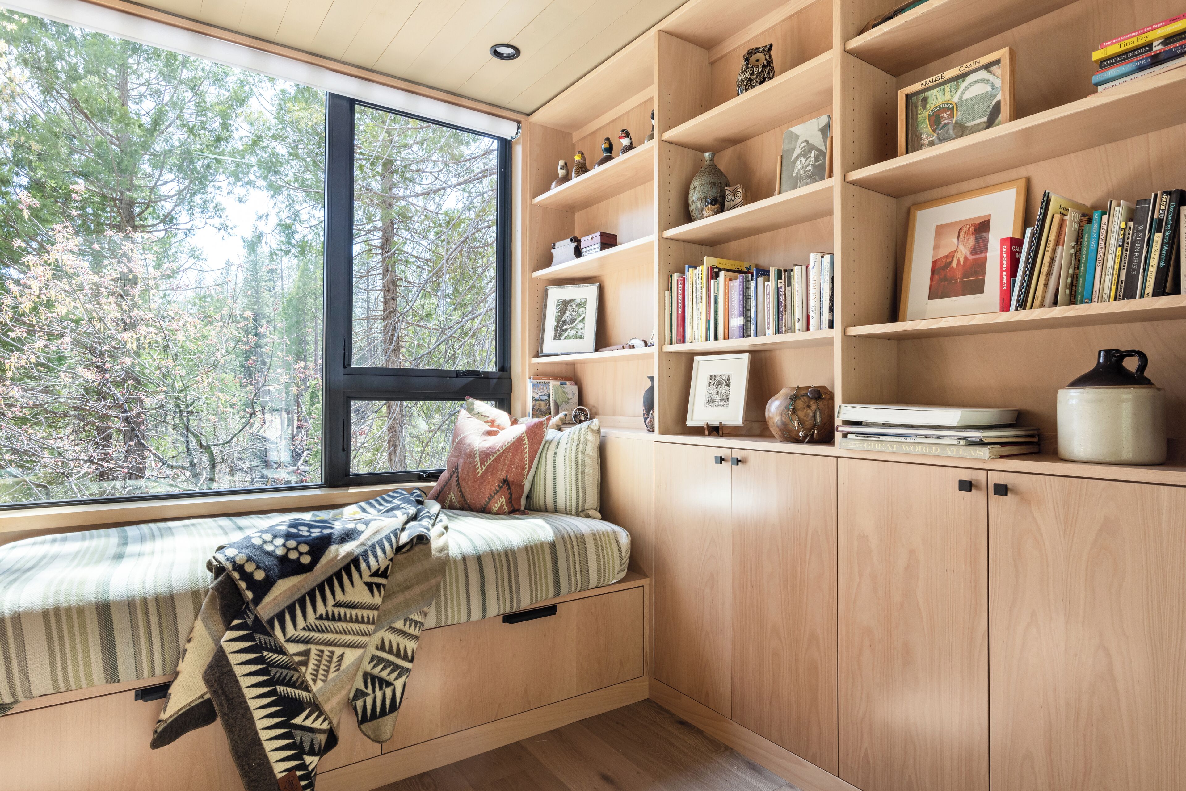 A cozy reading nook with a built-in bench, wooden shelves with books and decorative items, and a large window overlooking a forest.