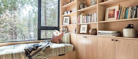 A cozy reading nook with a built-in bench, wooden shelves with books and decorative items, and a large window overlooking a forest.