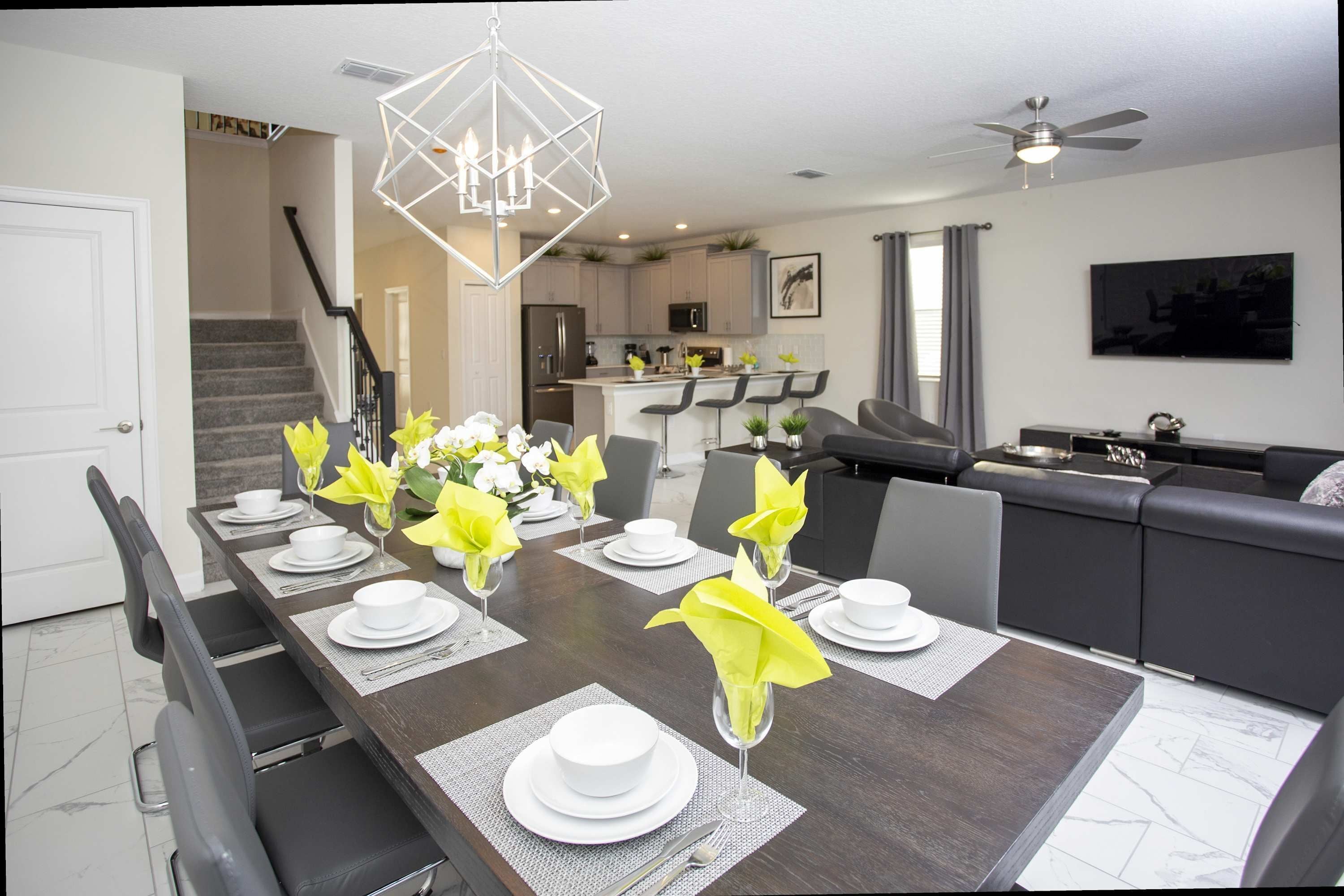 Een moderne eetruimte met een lange tafel gedekt voor zes personen, een keuken op de achtergrond en een trap die naar een andere verdieping leidt.