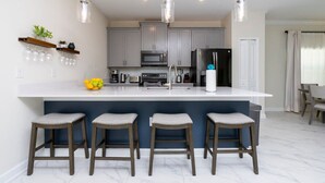 A modern kitchen with a central island, bar stools, pendant lighting, and a dining area with chairs. - Luxurious Family-friendly 5 Bd 12 Guests w Screened Pool Close to Disney (Kissimmee)
